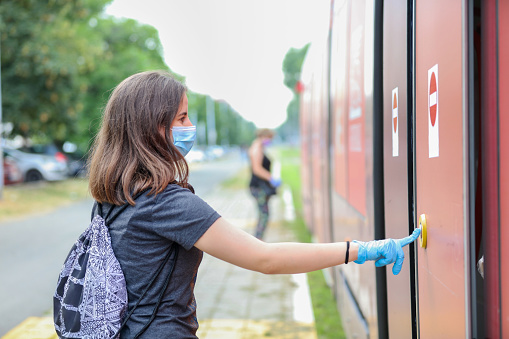 Teenage girl waiting for public transportation, pushing button on vehicle door in order to embark tramway, wearing protective face masks and surgical gloves in time of Covid-19 pandemia