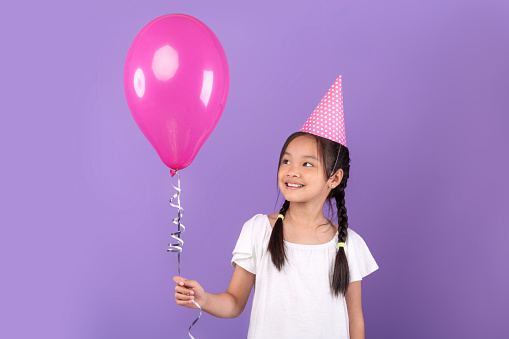 Smiling Korean Girl Wearing Pink Birthday Hat Holding Balloon Celebrating B-Day Standing Over Purple Background. Studio Shot