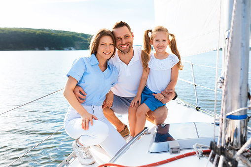 Joyful Family Of Three On Yacht Posing Sitting And Smiling To Camera Enjoying Sea Travel On Sailboat Outside. Boat Ride.