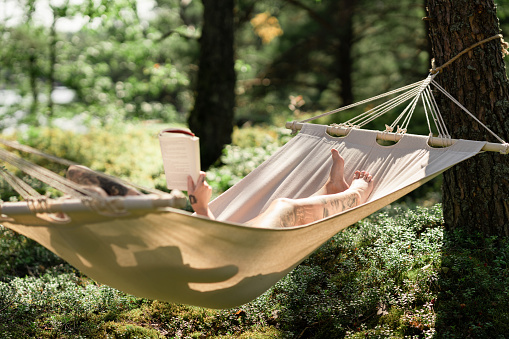 Woman reading a book on a sunny summer day. She is lying down in a hammock in a beautiful forest.