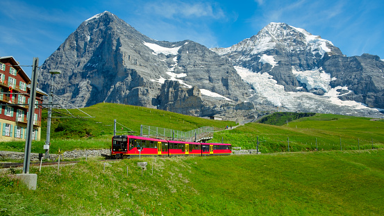 Kleine Scheidegg, Bernese Oberland, Switzerland - August 1 2019 : Red train from Jungfraujoch (Top of Europe) in fresh green alpine pastures. Eiger north face and monch in background