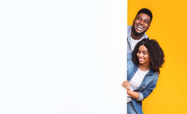 istock Free Space For Text. Cheerful african couple peeking from behind white board 1267294168