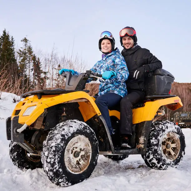 Portrait of romantic couple outdoor in winter in wild nature. Woman and man on yellow four-wheeler ATV in snowy mountains smiling at camera. Concept of relationships, active leisure, winter activities