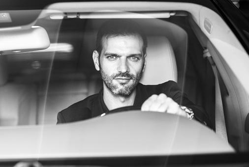 Cool man in his 30s sitting behind the steering wheel driving. He is wearing a suit and staring at the camera. Black and white.