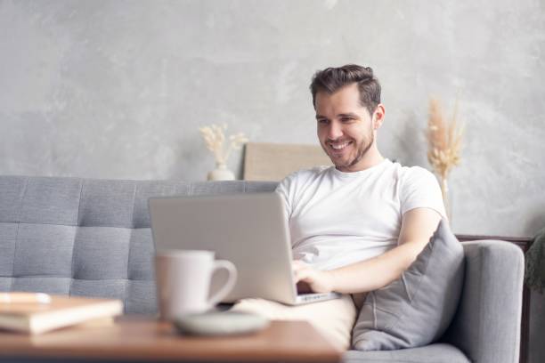 istock Typing new blog post. Handsome young man using his laptop with smile while sitting on the couch at home. 1267317306