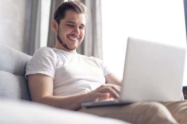 istock Typing new blog post. Handsome young man using his laptop with smile while sitting on the couch at home. 1267317311