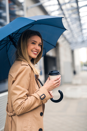 Charming young lady walking down the street holding umbrella and cup of coffee, smiling at the camera
