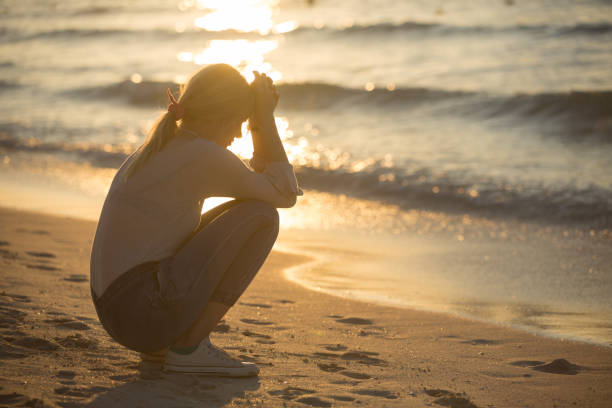 Sad woman alone at the beach during sunset. Love, loneliness and heartbreak. Heart broken young female person suffering alone after a breakup. She is sitting on the sand near the sea water in the evening while the sun sets. crying photos stock pictures, royalty-free photos & images