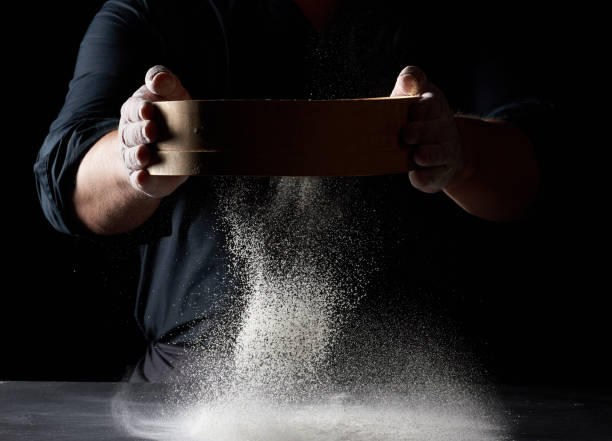 chef un hombre con un uniforme negro sostiene un tamiz redondo de madera en sus manos y tamiza la harina de trigo blanco sobre un fondo negro - sifting fotografías e imágenes de stock