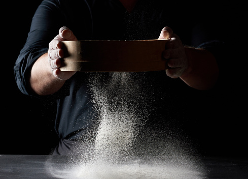 chef a man in a black uniform holds a round wooden sieve in his hands and sifts white wheat flour on a black background, the particles fly in different directions