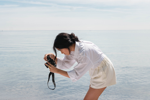 An Asian woman takes a picture, using her camera, while on vacation at the beach.