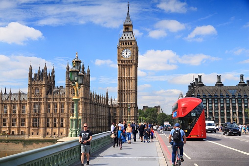 View across River Thames of Palace of Westminster, Elizabeth Tower with landmark clock face, and commuters crossing green arch bridge under dramatic summer sky.