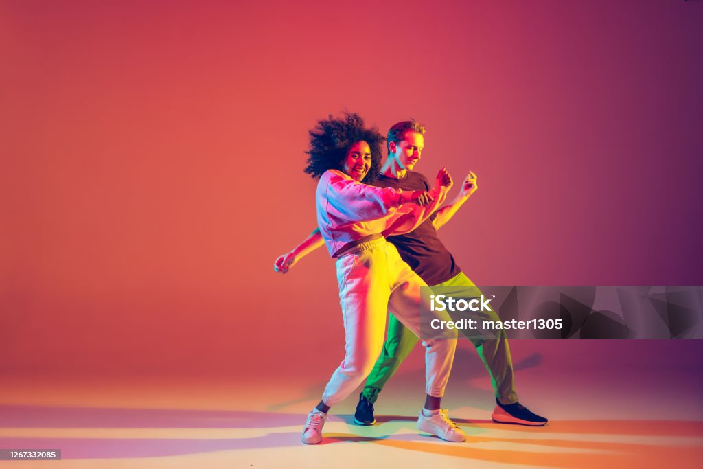 Stylish man and woman dancing hip-hop in bright clothes on gradient background at dance hall in neon light Drive in motion. Stylish man and woman dancing hip-hop in bright clothes on green background at dance hall in neon light. Youth culture, movement, style and fashion, action. Fashionable portrait. Dancing Stock Photo