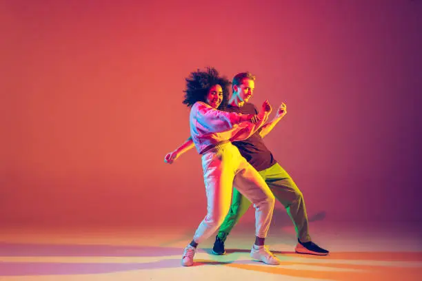 Drive in motion. Stylish man and woman dancing hip-hop in bright clothes on green background at dance hall in neon light. Youth culture, movement, style and fashion, action. Fashionable portrait.