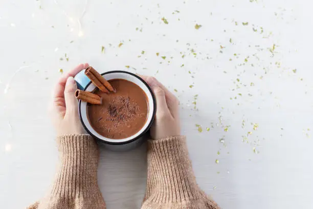 Photo of Hot chocolate with cinnamon spice in enamel mug in female hands on white wooden background top view.