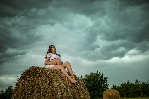 A beautiful young pregnant woman sitting at hay bale at  stubble field and enjoying the last days of pregnancy. She enjoying the beautiful landscape and nature around her while she holding and cuddling her pregnant belly. The weather is cloudy and windy with very dramatic sky