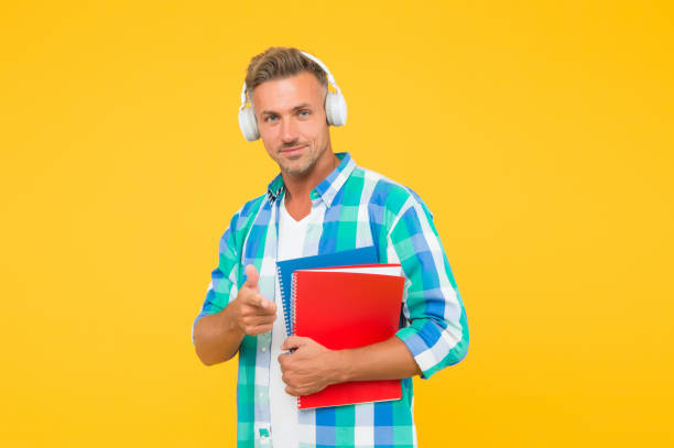 istock handsome university tutor pointing finger. adult student hold notebook. man with folder in headphones. online education. man hold book for notes while listen ebook. lets write in notepad 1267339302