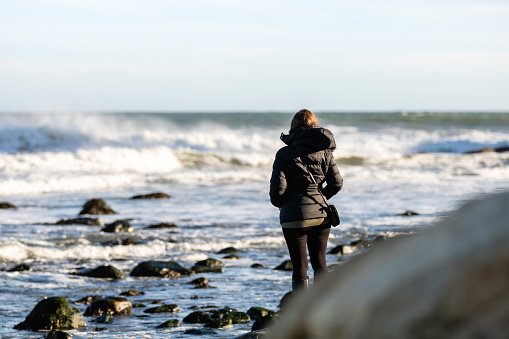 Montauk, New York - January 1, 2019 : Young woman in a black coat visiting the beach, gazing off into the distance at the rough sea.