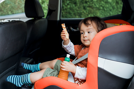 A young boy sitting in a car seat with a snack and drink in casual summery clothing during lockdown.