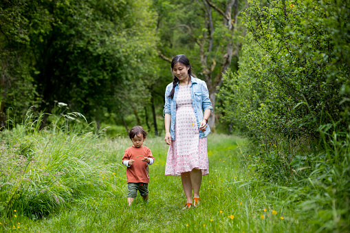 A young boy and his pregnant mother in casual summery clothing. They are smiling and walking through a forest during lockdown.