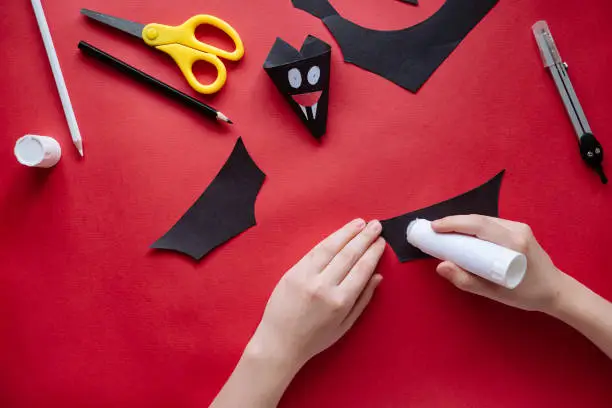 Photo of How to make bat out of paper at home. Hands making craft out of paper. Step by step photo instruction. Step 10. Glue the wings. Children DIY art project.