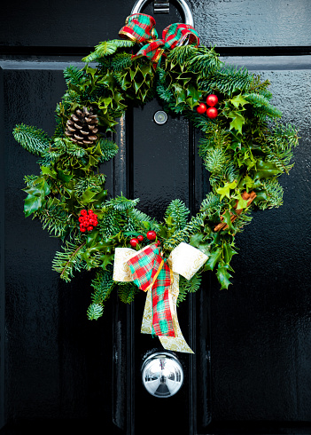 A traditional decorative yule Christmas wreath made of pine branches, pine cones, dried oranges, cinnamon bun and red berries hanging on a hanging on a black colour wooden Victorian front door with  knocker in London, UK