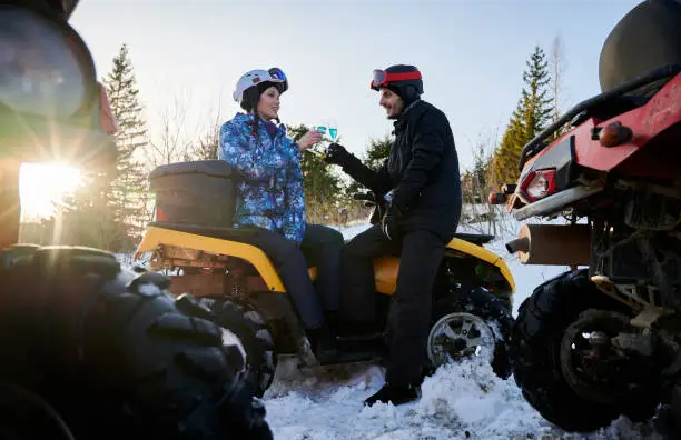Young couple wearing winter ski suits and helmets, sitting on yellow ATV holding two glasses with blue champagne looking at each other. Sunny winter day. Huge wheels of other ATV are on foreground