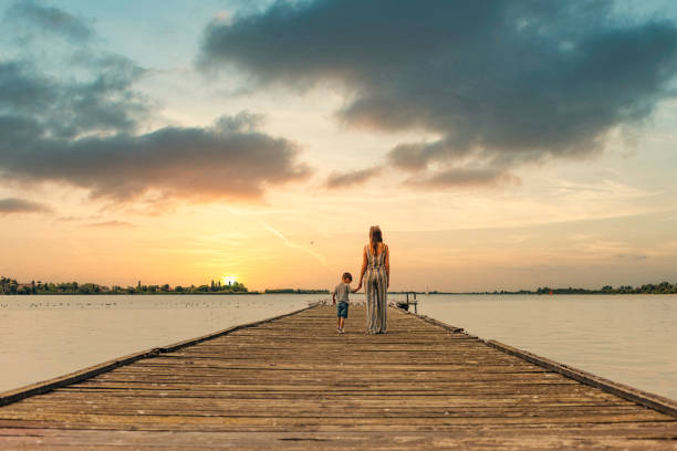 Happy single mother and her three years old son relaxing on a wooden dock lake side during sunset.