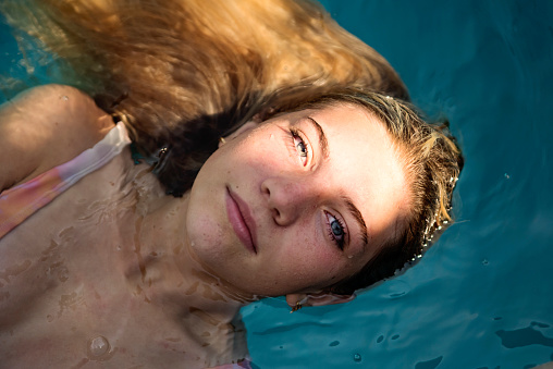 Beautiful teenage girl looking at camera for a portrait in pool backyard on a very sunny summer day. She has long hair and is wearing a swimsuit. Horizontal waist up outdoors shot with copy space.