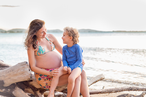 Toddler sitting on the lap of his pregnant mother at the beach. Looking at each other face to face.