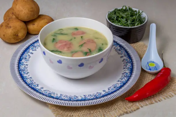 Caldo Verde Soup is a popular soup in Portuguese cuisine with greens and chopped chorizo on the top in ceramic bowl and piece of bread and pepper. Top View