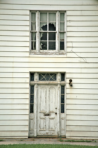 Old abandoned weathered wooden house entrance with textured door & window with torn curtain