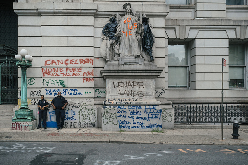 Manhattan, New York. July 07, 2020. A police woman and a police man wearing a mask stand at the Surrogate Hall of Records entrance in Lower Manhattan next to graffitis and the vandalized statue by Philip Martiny.