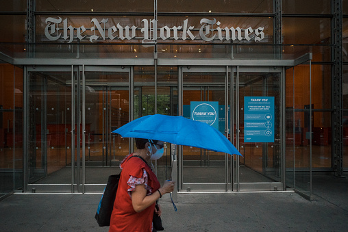 Manhattan, New York. July 10, 2020. A woman wearing a mask with an umbrella walks in front of the main entrance of the New York Times building in Midtown.