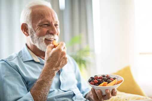 Happy senior man eating healthy fruits while relaxing on sofa at home.