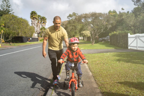 Father supporting son to lean bike. Father helping his son to learn bike ride outside of house in the street on pathway. auckland region photos stock pictures, royalty-free photos & images