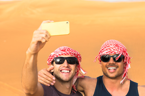Tourism. Cheerful young guys smiling while taking a picture with the golden Arabian desert sand dunes in the background. They are enjoying a desert safari tour while wearing the local headwear and sunglasses.