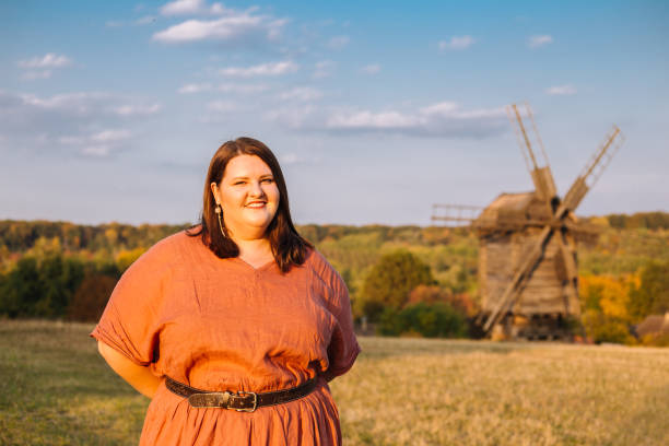 istock Beautiful and confident plus size model in red dress and brown boots posing outdoors at rural autumn landscape with wooden mill 1267456561