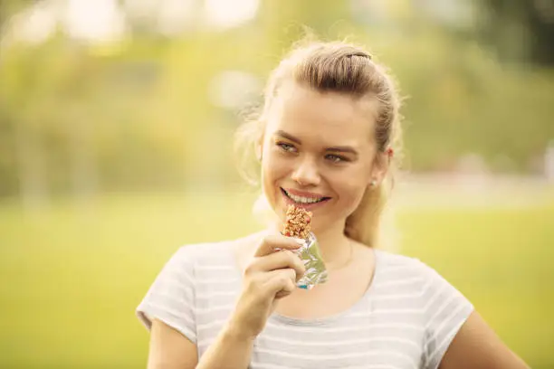 Photo of Sportive Woman eating a protein bar after outdoor workout - Closeup face of young blonde sporty woman resting while biting a nutritive bar
