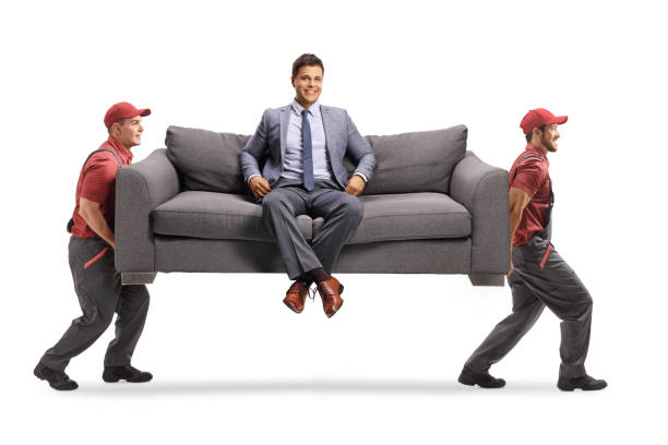 istock Smiling man in elegant clothes sitting on a sofa at home and two movers carrying the sofa 1267466885