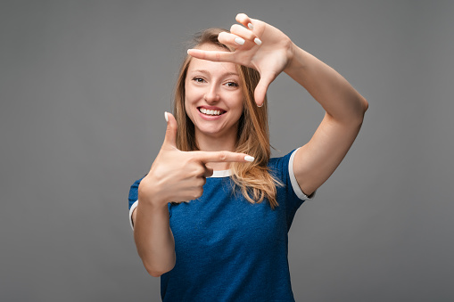 Portrait of positive young blonde female with cheerful expression, dressed in casual blue t shirt, has good mood, gestures finger frame actively at camera. Human emotions, facial expression concept
