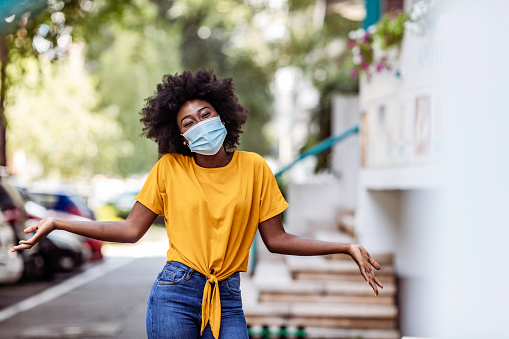 Delightful African-American woman wearing surgical protective mask  standing on empty sunny city street during COVID-19 quarantine while looking at camera with smile.