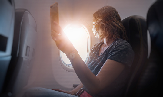 Traveling by airplane during coronavirus pandemic. Woman with face mask near porthole, looking through the airplane window