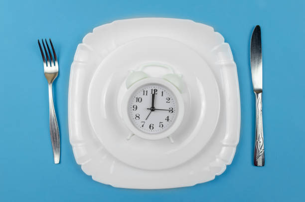 The Top Ten Health Benefits of Fasting: From Weight Loss to Longevity 4 white alarm clock is on a white round plate which is in a larger white plate with a knife on