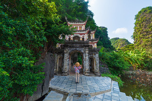 Lone tourist with traditional vietnamese hat at Bich Dong Pagoda entrance gate, Ninh Binh Vietnam, buddhist temple set amid jungle and karst mountain range. Traveling alone, keep social distancing.