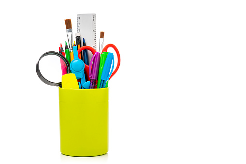 Front view of a green desk organizer filled with colorful office or school supplies isolated on white background. The composition is at the left of an horizontal frame leaving useful copy space for text and/or logo at the right. High resolution 42Mp studio digital capture taken with Sony A7rII and Sony FE 90mm f2.8 macro G OSS lens