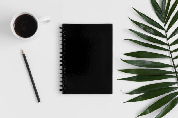 istock Top view of a black notebook mockup with  workspace accessories and a palm leaf on a white table. 1267490818