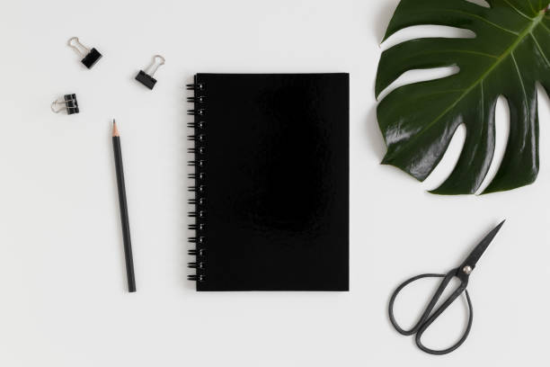 istock Top view of a black notebook mockup with  workspace accessories and a monstera leaf on a white table. 1267490857