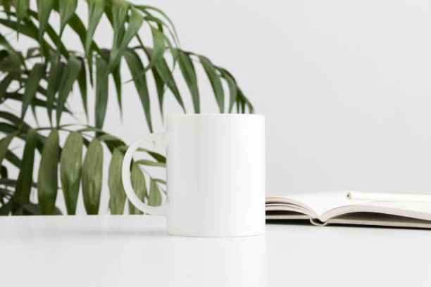 istock Mug mockup with notebook on a white table and a palm leaf. 1267495290