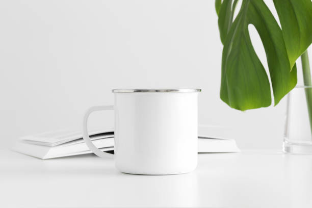 istock Enamel mug mockup with a monstera leaf in a vase and a book on a white table. 1267500989